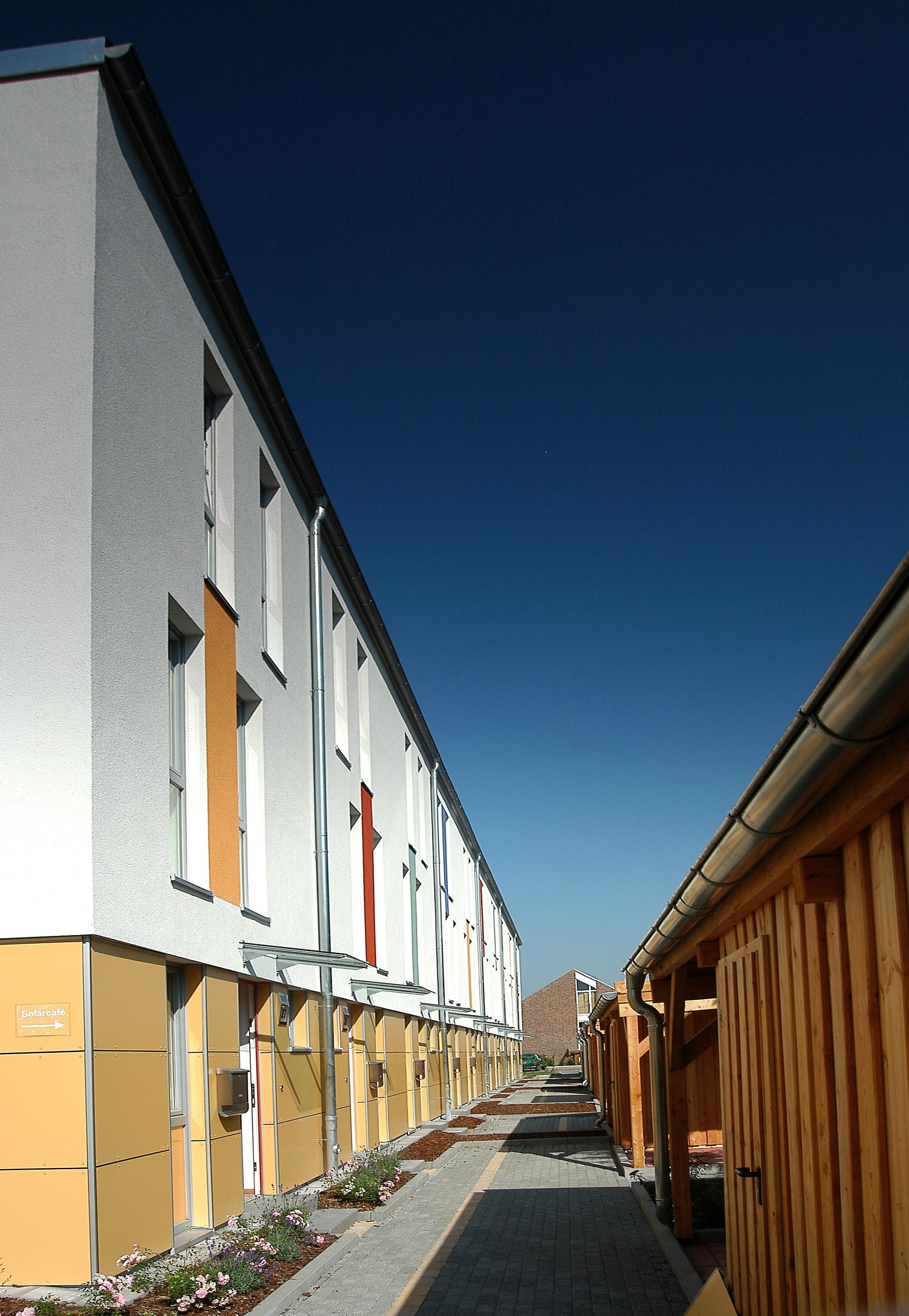 Eine schmale Gasse zwischen weißen Wohnhäusern und Holzschuppen unter klarem, blauem Himmel