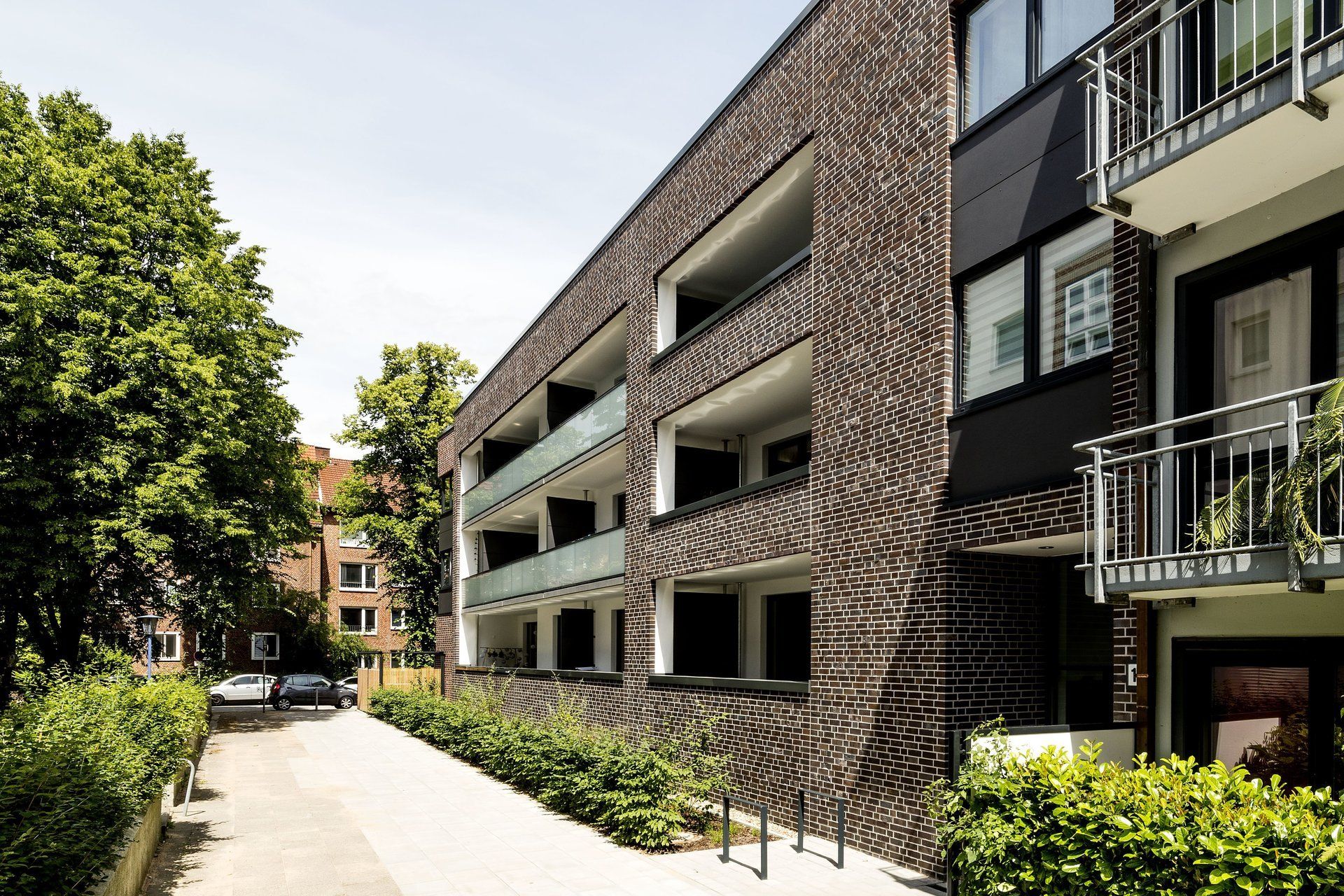 Modernes Mehrfamilienhaus mit Balkonen entlang eines sonnigen Gehwegs, der von Bäumen und Sträuchern gesäumt ist.