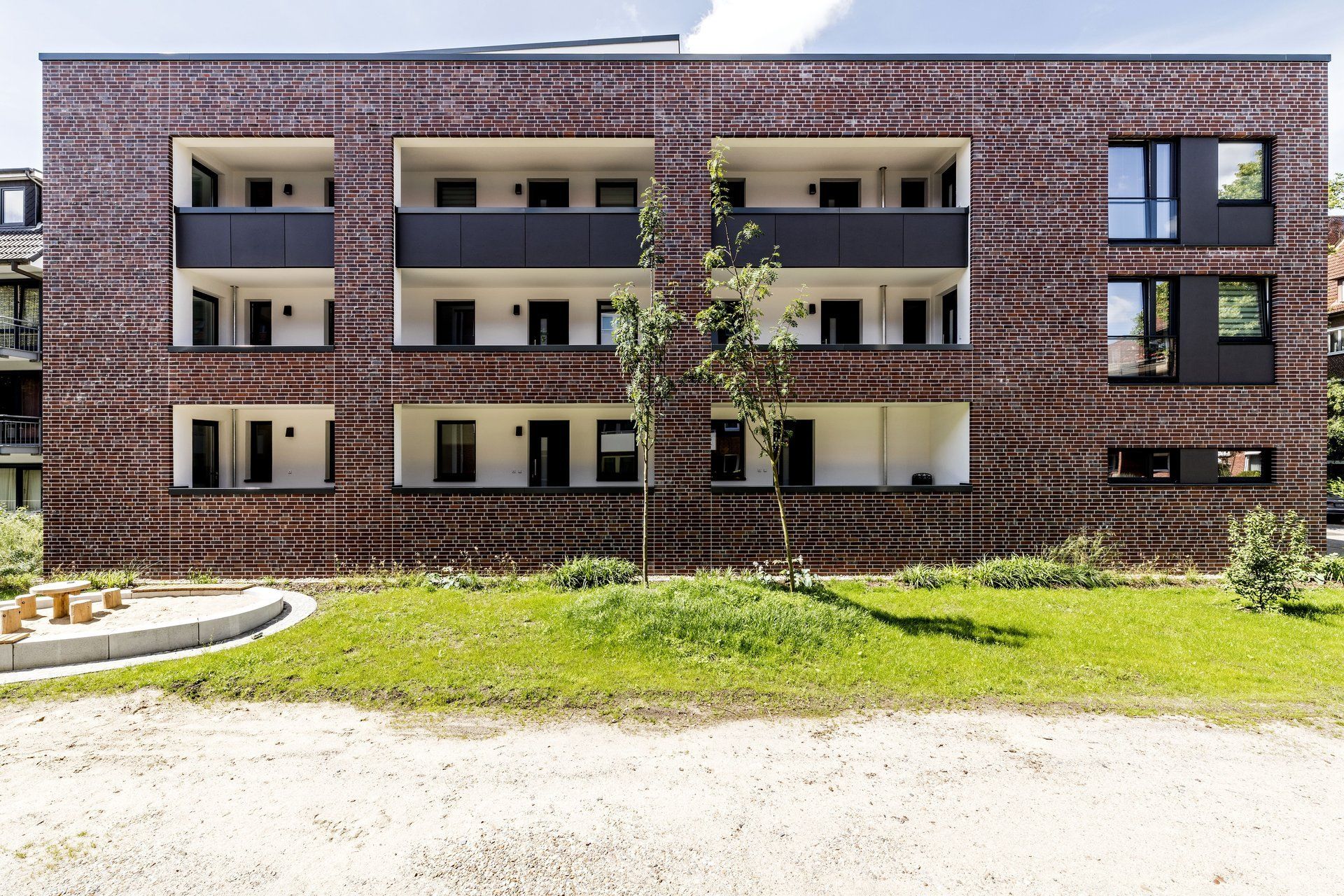 Mehrfamilienhaus aus Backstein mit drei Balkonen, Fenstern und einem begrünten Vorgarten.
