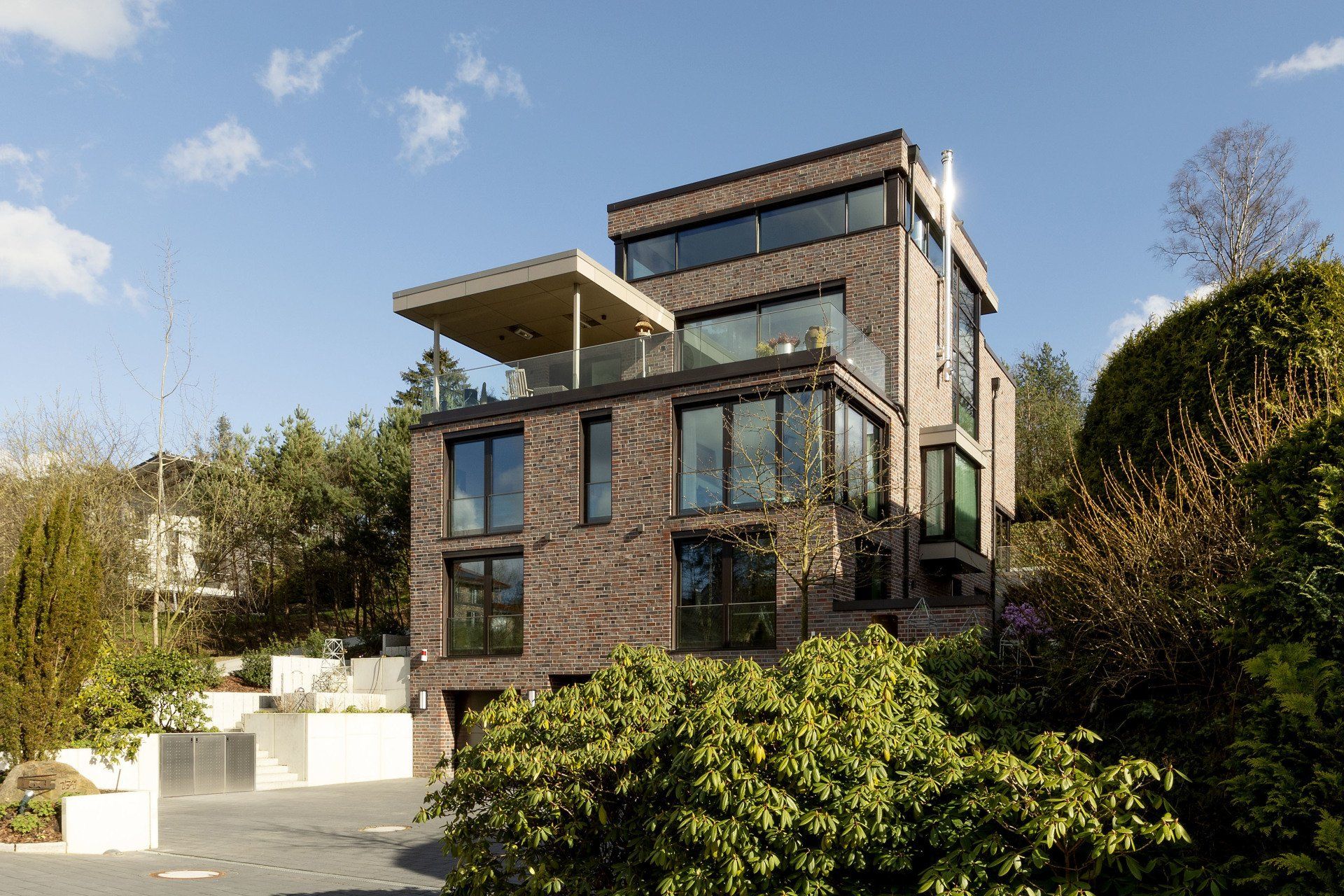 Modernes Backsteinhaus mit großen Fenstern und Dachterrasse inmitten üppigen Grüns unter blauem Himmel