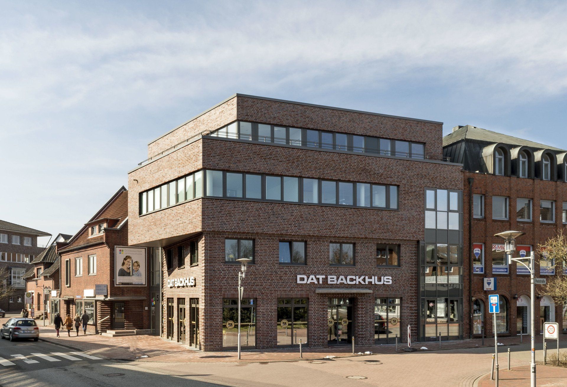 Modernes Eckgebäude aus Backstein mit großen Fenstern und Ladenlokal im Erdgeschoss an einer sonnigen Straße.