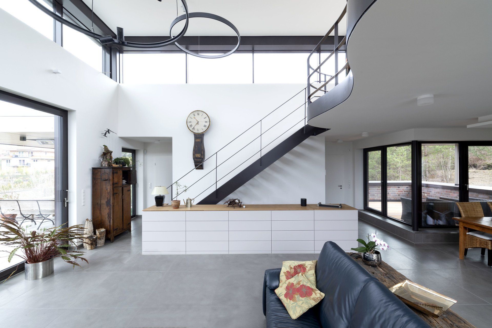 Modernes, offenes Wohnzimmer mit Treppe, weißen Wänden, hoher Decke und dunklem Sofa im Vordergrund