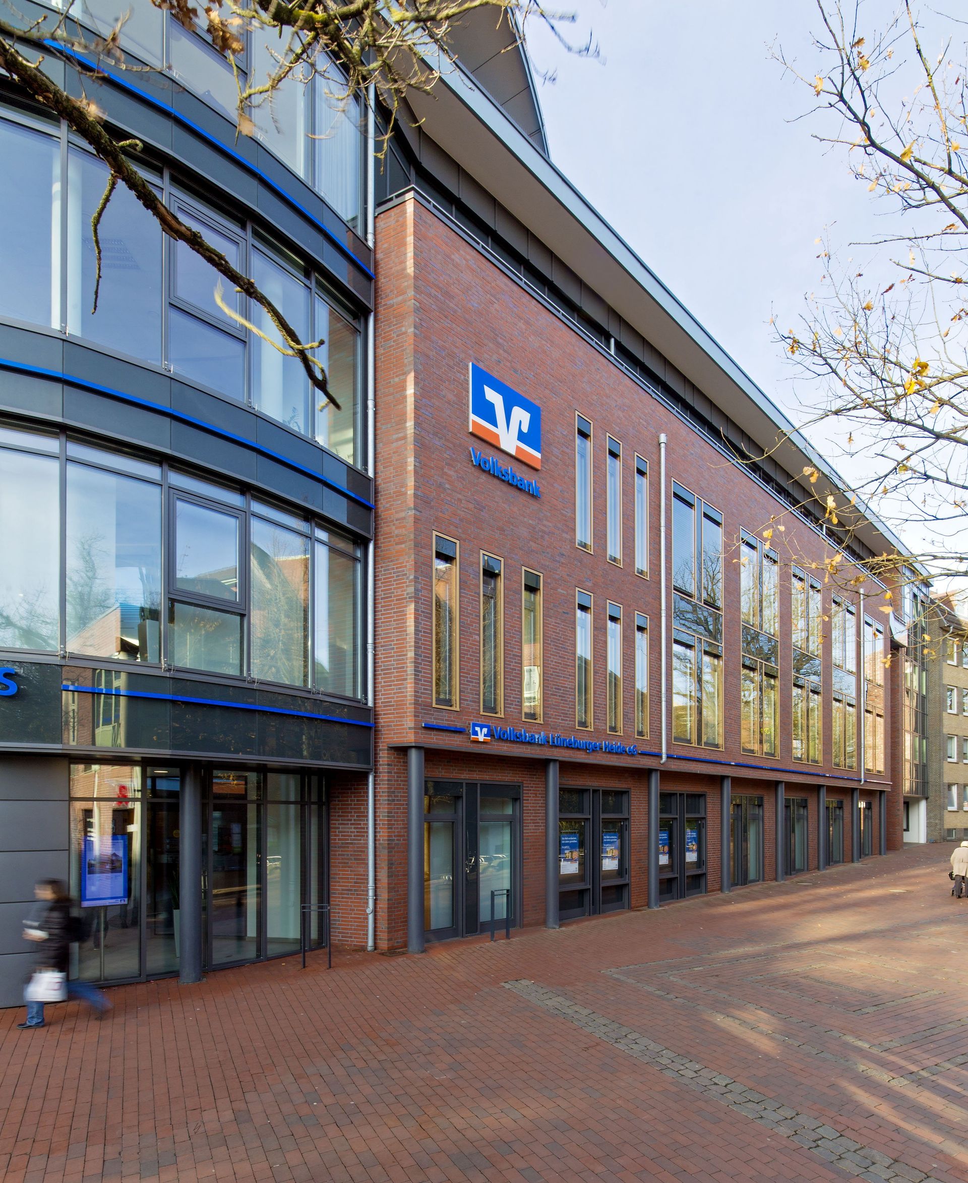 Straßenansicht eines modernen Geschäftsgebäudes aus Backstein und Glas in einer gepflasterten Fußgängerzone.