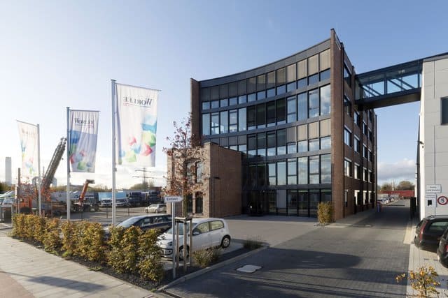 Modernes Bürogebäude mit großen Fenstern, geparkten Autos und Fahnen draußen an einem sonnigen Tag