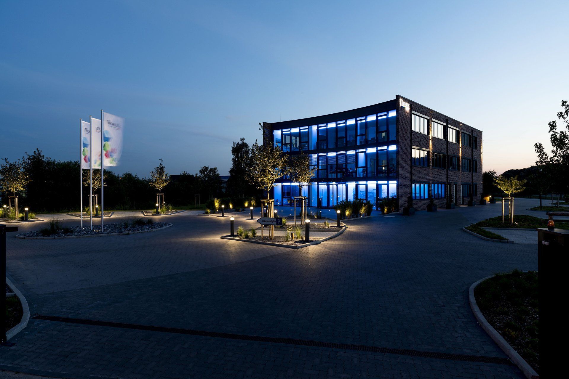 Gebäude in der Abenddämmerung mit blau beleuchteten Fenstern, einem gepflasterten Innenhof und Fahnenmasten im Außenbereich.