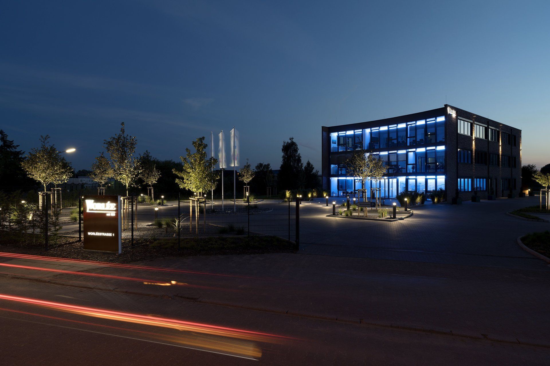 Beleuchtetes modernes Bürogebäude bei Nacht mit blau beleuchteten Fenstern und Scheinwerferlichtspuren von Autos davor