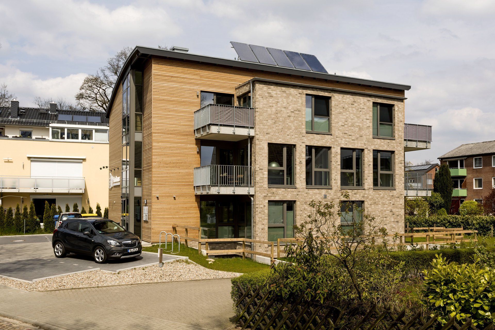 Modernes dreistöckiges Mehrfamilienhaus mit Holz- und Steinfassade, Solaranlage und davor geparktem Auto.