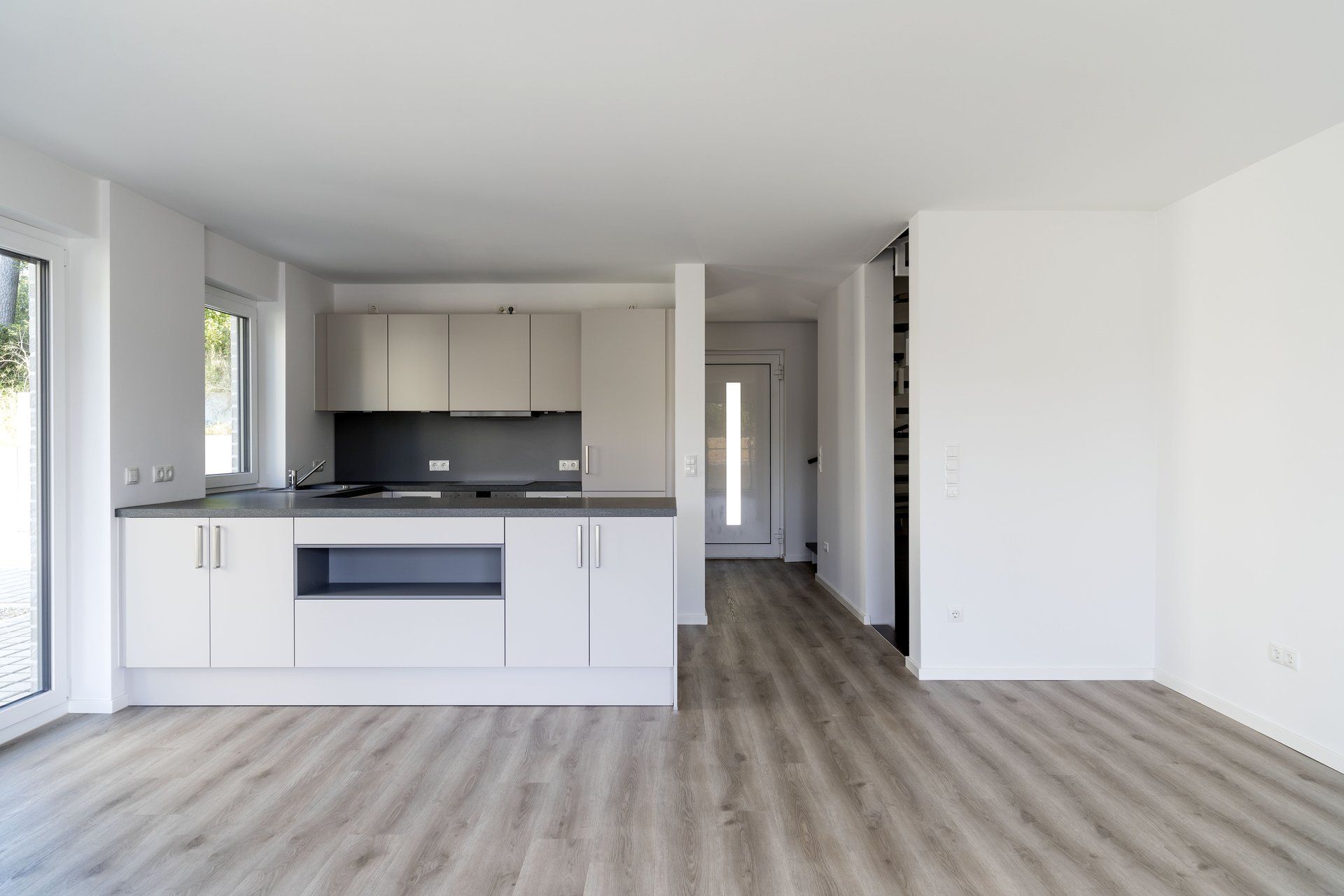 Helle, leere, moderne Küche mit weißen Schränken, schwarzem Fliesenspiegel und grauem Holzboden