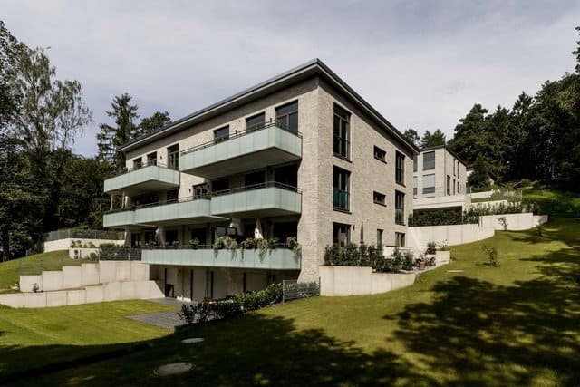 Modernes Steinhaus mit Balkonen an einem grasbewachsenen Hang, umgeben von Bäumen