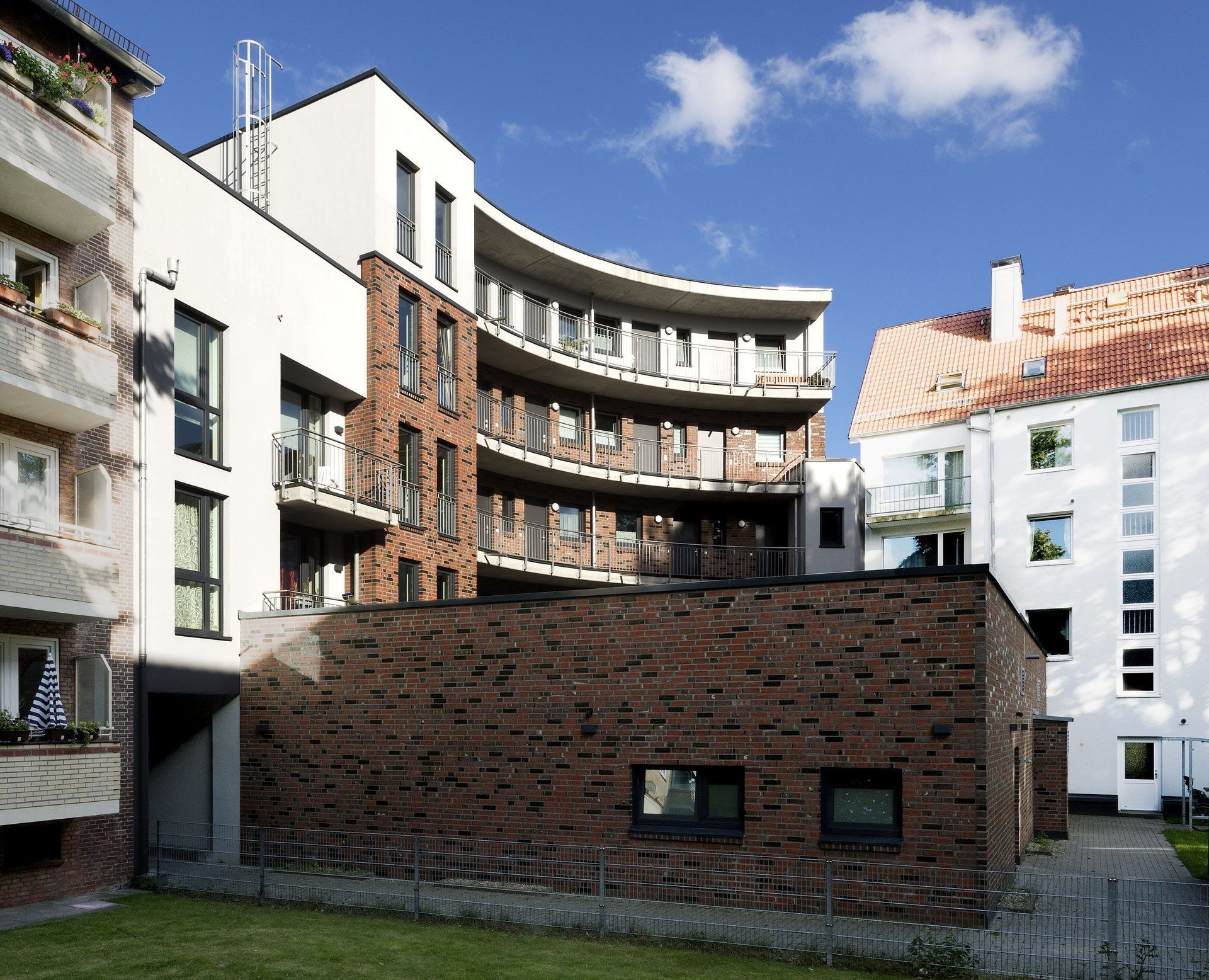 Moderner Apartmenthof mit geschwungenen Balkonen, Backsteinmauern und einer Rasenfläche unter blauem Himmel