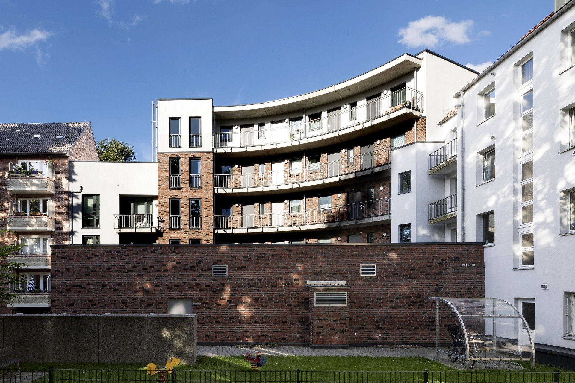 Modernes Apartmentgebäude mit geschwungenen Balkonen hinter einer Backsteinmauer und grünem Rasen unter blauem Himmel