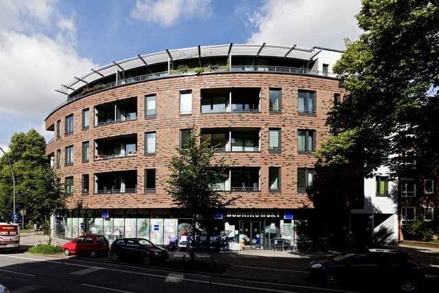 Geschwungenes Backstein-Wohnhaus mit Geschäften im Erdgeschoss, geparkten Autos und Bäumen an einer Stadtstraße.