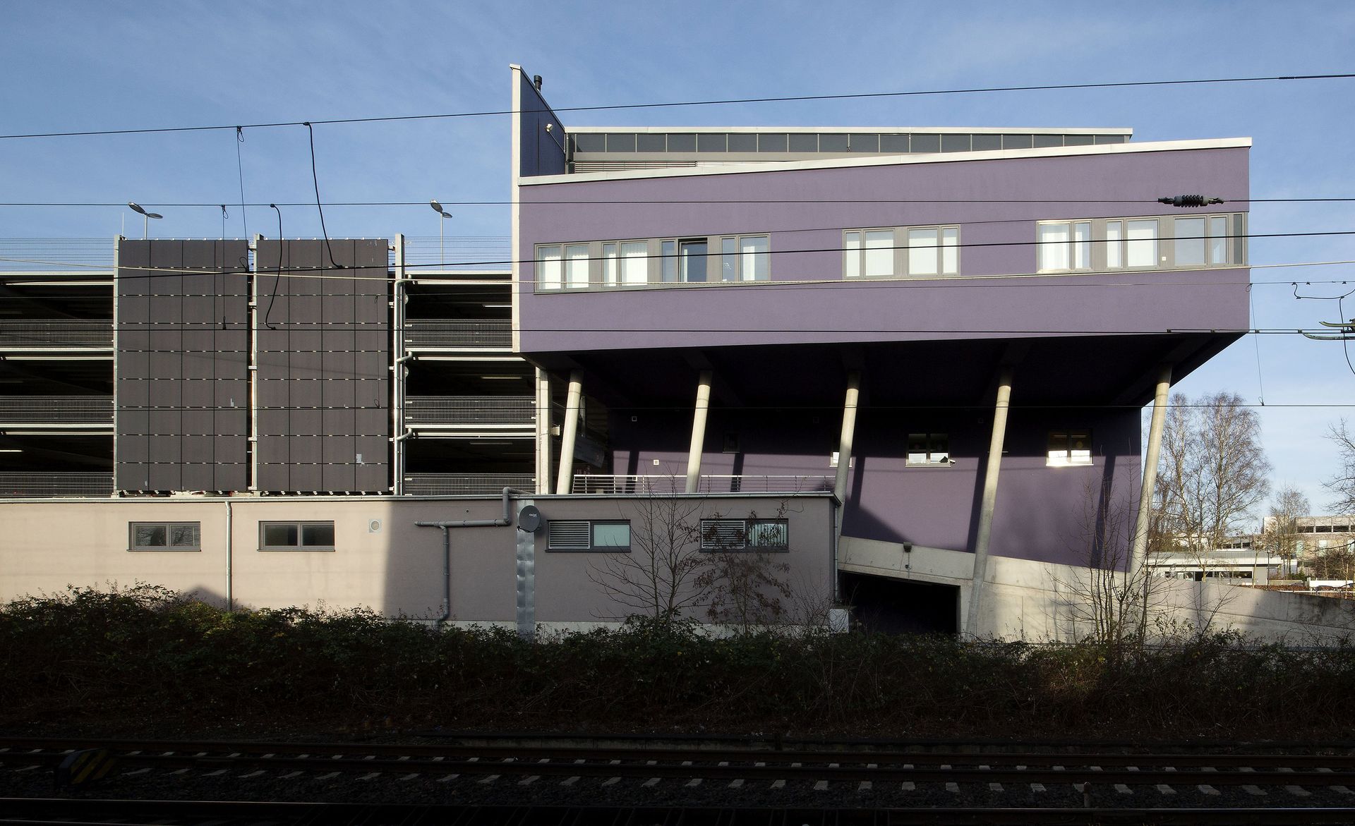 Modernes, lila-graues Gebäude mit Balkonen und Parkhaus hinter einer Hecke
