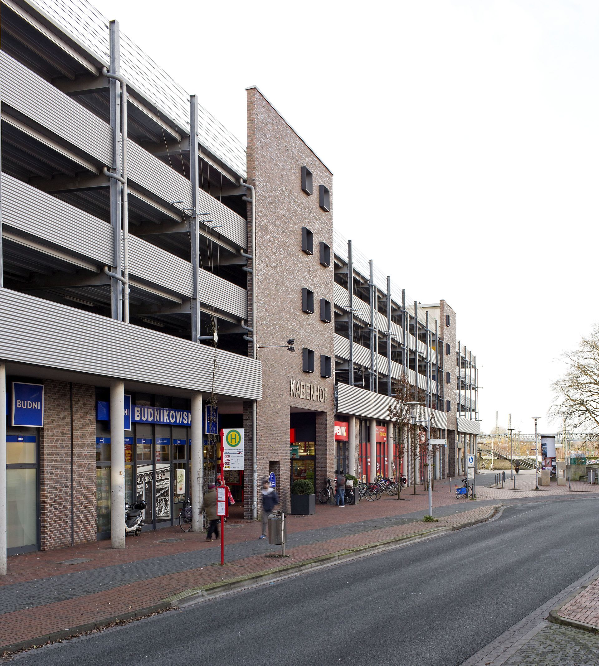 Eine Reihe von Backsteinfassaden neben einer Schotterstraße, mit geparkten Fahrrädern und einem mehrstöckigen Parkhaus.