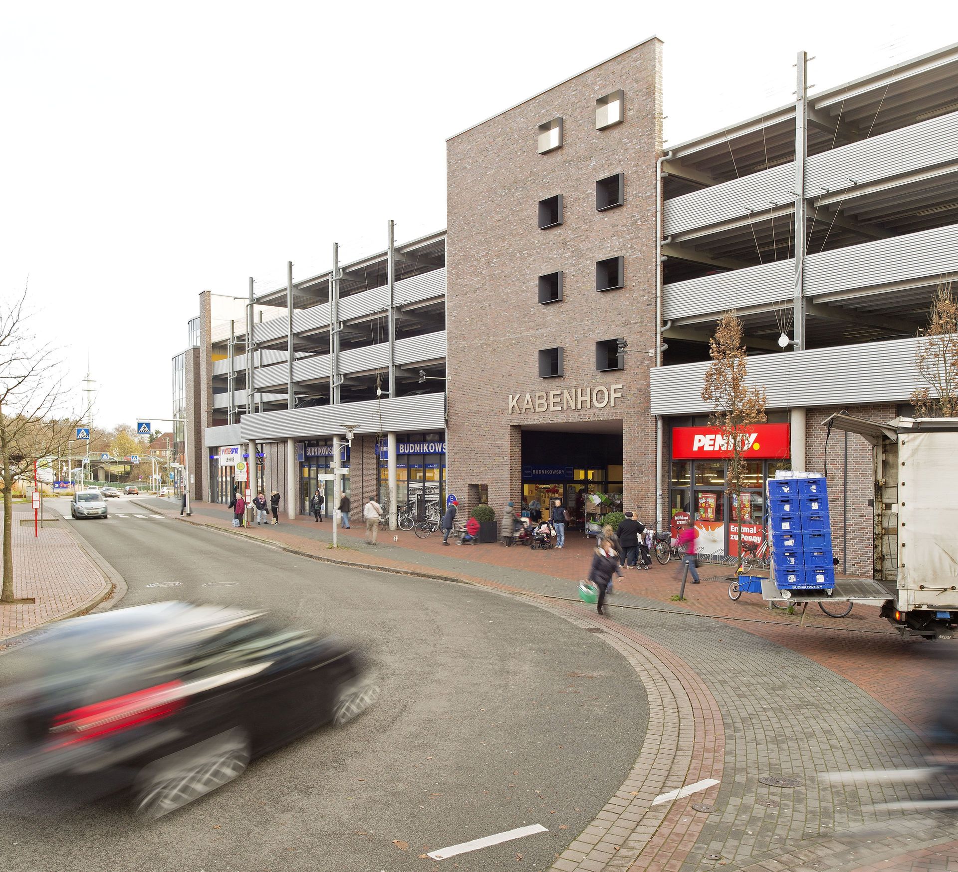 Kurvenreiche Straße neben einem gemauerten Parkhaus, im Vordergrund ein verschwommenes Auto, das vorbeifährt.