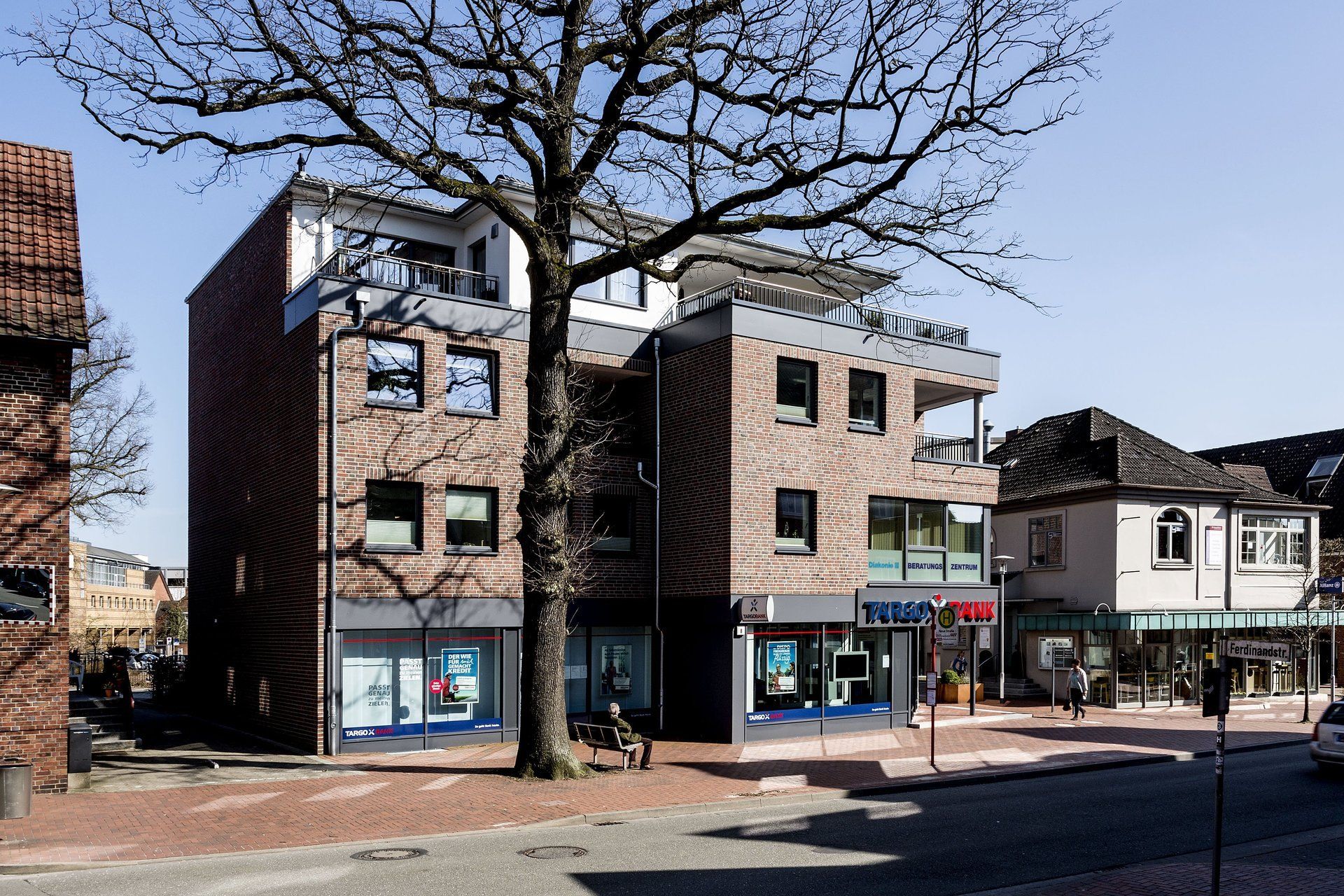 Straßenansicht von Backsteingebäuden mit Ladenfronten und kahlen Bäumen an einem sonnigen Tag