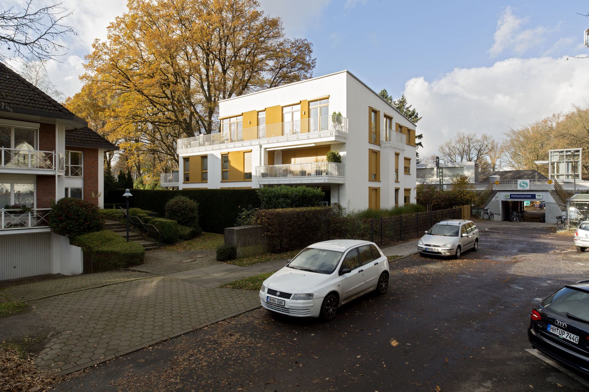Ruhige Wohnstraße mit geparkten Autos und einem modernen weißen Wohnhaus unter herbstlichen Bäumen