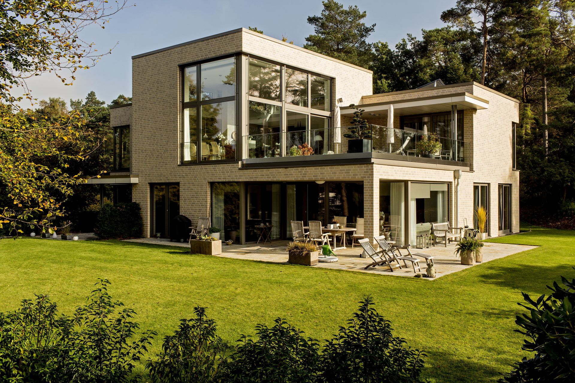 Modernes zweistöckiges Haus mit großen Glasfenstern, Terrasse und grünem Rasen in einem sonnigen Garten.