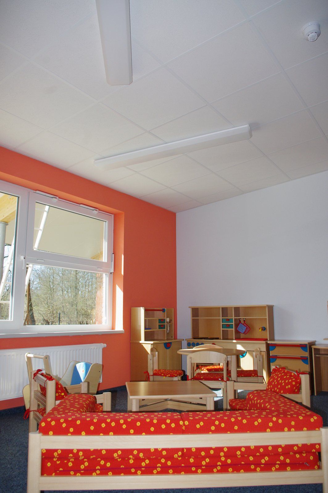 Farbenfrohes Klassenzimmer mit roten Betten, Regalen und einem großen Fenster unter einer weißen Decke