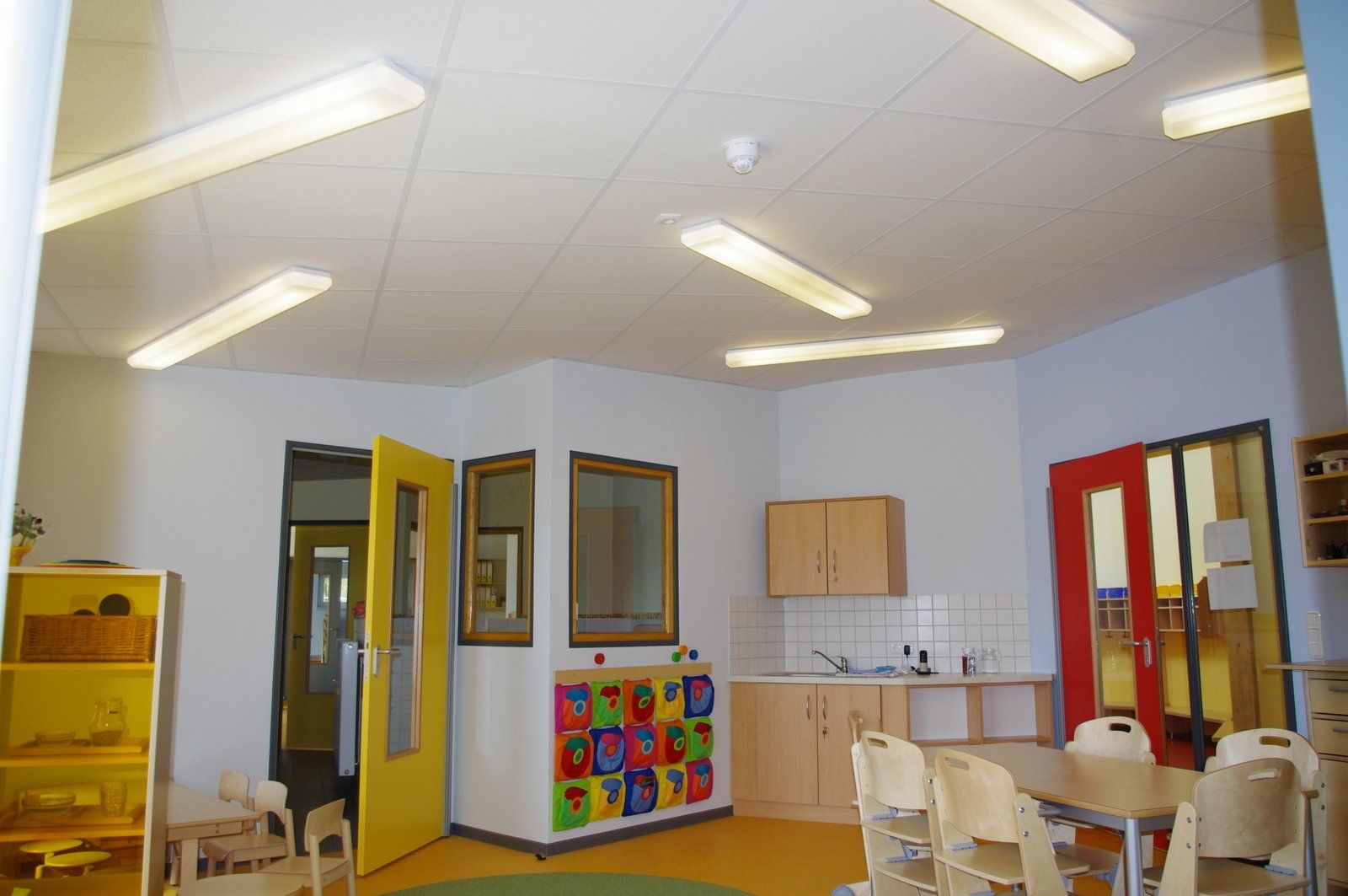 Helles Vorschulklassenzimmer mit Tischen, Stühlen, farbenfrohen Fächern und großen Fenstern.