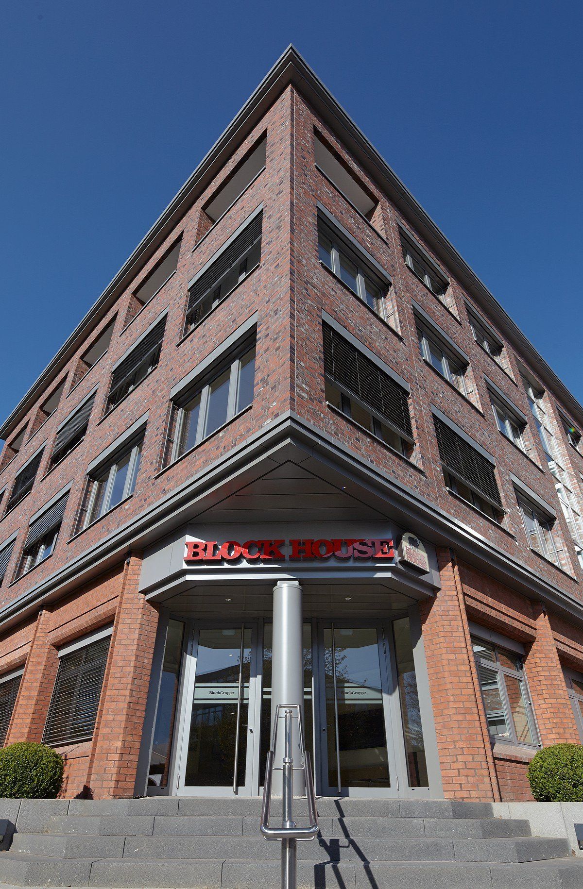 Eingang eines Backsteingebäudes mit roter Markise und Glastüren, von unten vor blauem Himmel gesehen