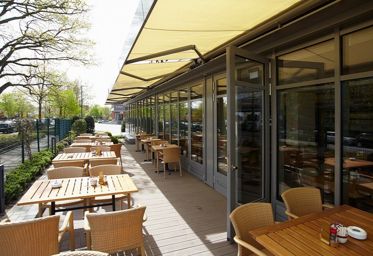 Außenterrasse eines Cafés mit Holztischen und -stühlen unter einer gelben Markise neben Glasfenstern