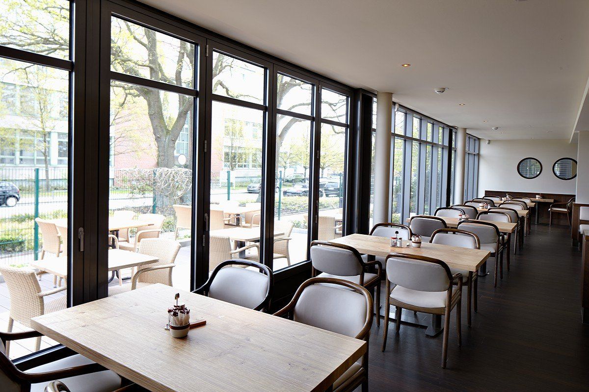 Leeres, modernes Café mit großen Fenstern, weißen Tischen und Stühlen und Blick auf die Bäume draußen.