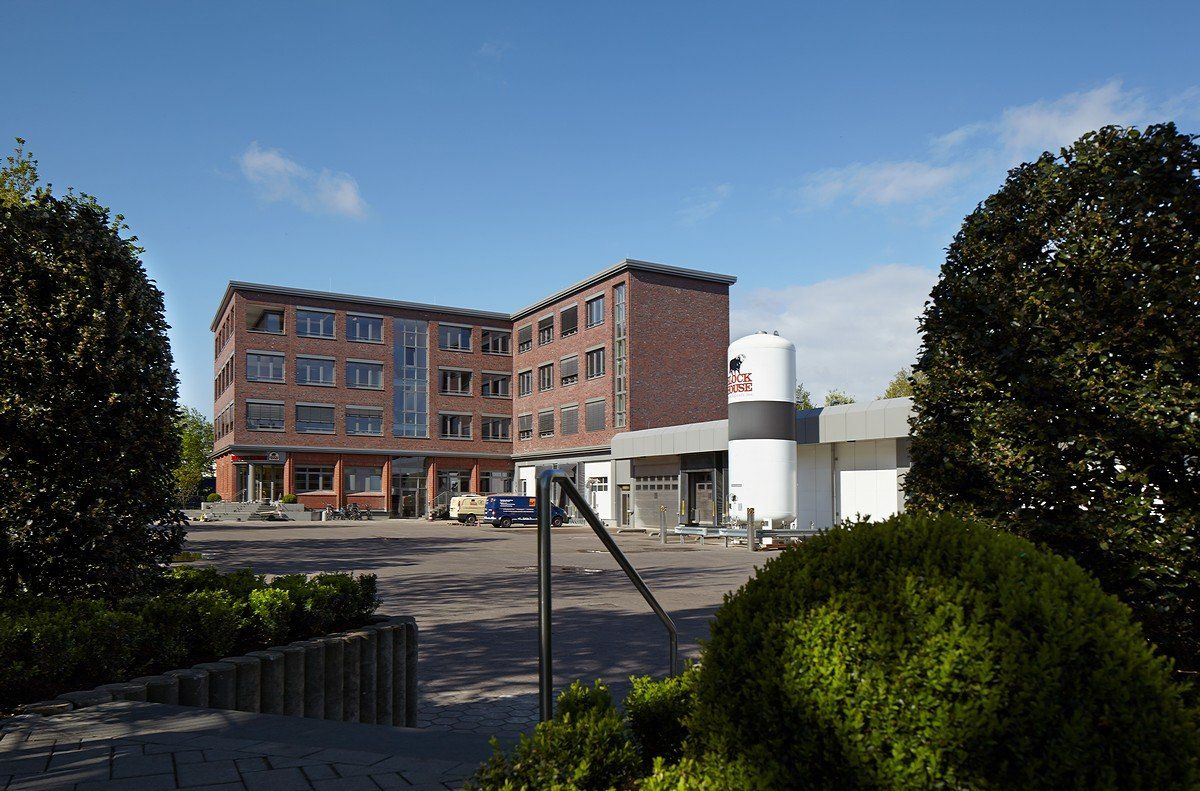 Backsteingebäude und weiße, siloartige Struktur hinter Sträuchern in einem sonnigen Innenhof unter blauem Himmel