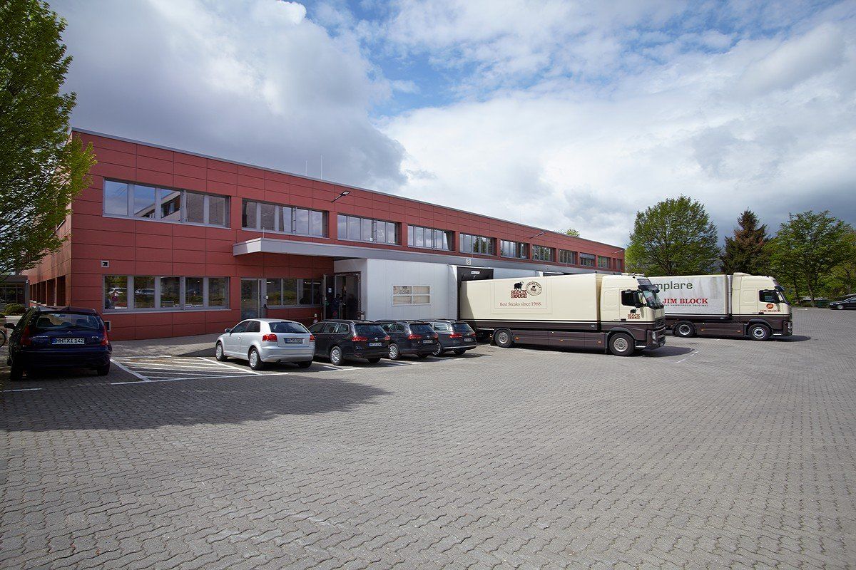 Rotes Industriegebäude mit geparkten Autos und Lieferwagen auf einem gepflasterten Parkplatz unter bewölktem Himmel