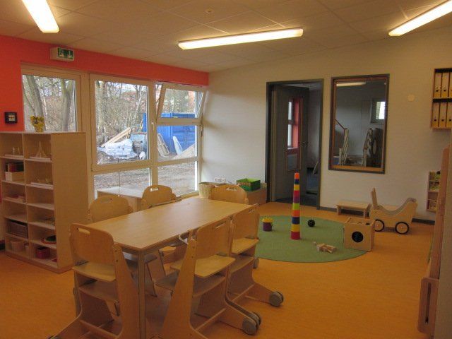 Ruhiges Klassenzimmer mit kleinen Tischen, Stühlen, Regalen und einer Spielecke in der Nähe großer Fenster.