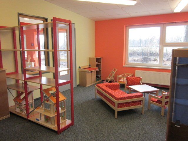 Farbenfrohes Klassenzimmer mit roten Regalen, kleinen Tischen und einem Bett an einem großen Fenster