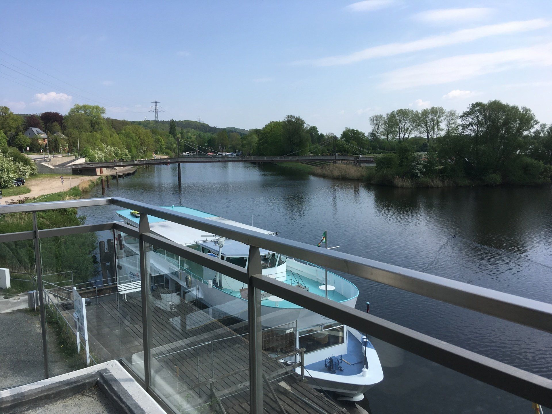 Flussblick vom Balkon mit Bootsanleger, ruhigem Wasser und Bäumen unter klarem Himmel.