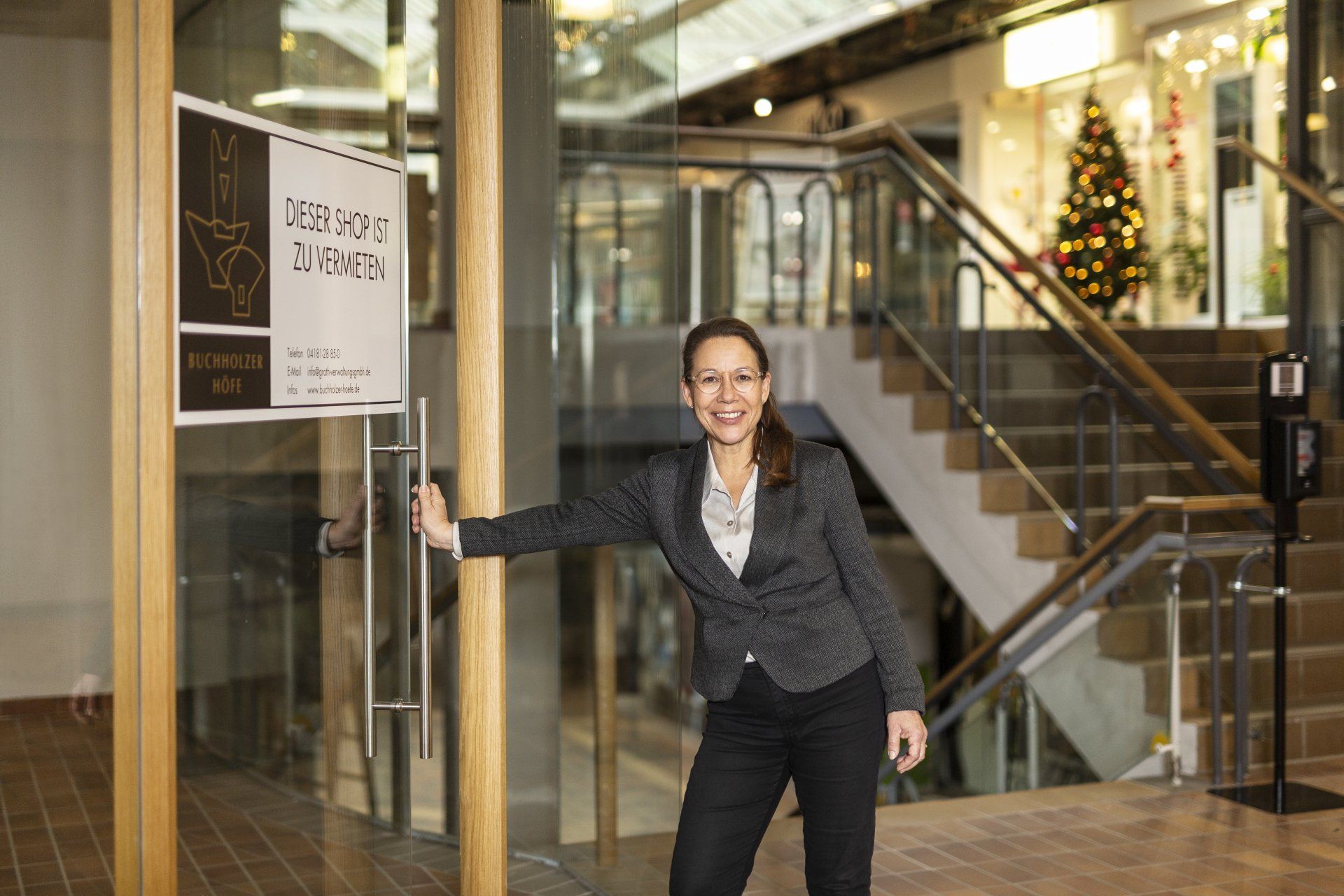Eine Frau öffnet eine Glastür in einer modernen Lobby mit Treppe und einem geschmückten Weihnachtsbaum.