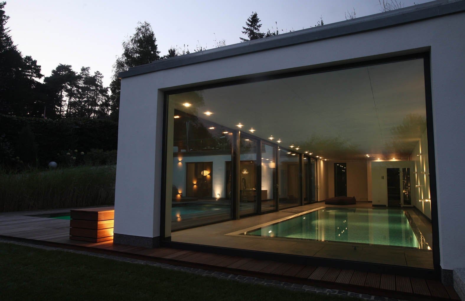 Modernes Haus mit großer Glaswand, die bei Dämmerung einen beleuchteten Innenpool freigibt.