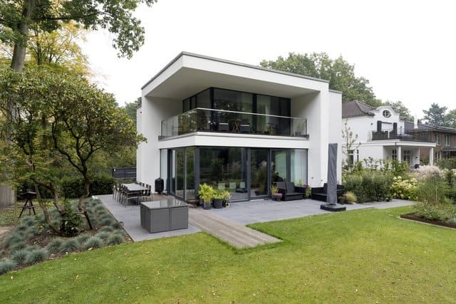 Modernes, zweistöckiges weißes Haus mit großen Glasfenstern, Terrasse und grünem Rasen, umgeben von Bäumen