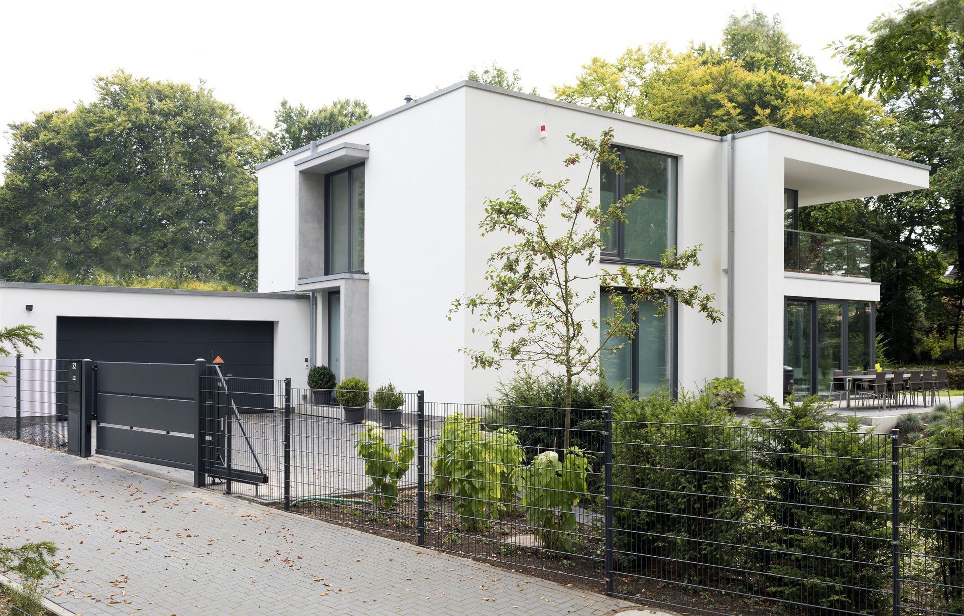 Modernes weißes Haus mit schwarzer Garage und schwarzem Zaun, umgeben von Bäumen und Grün