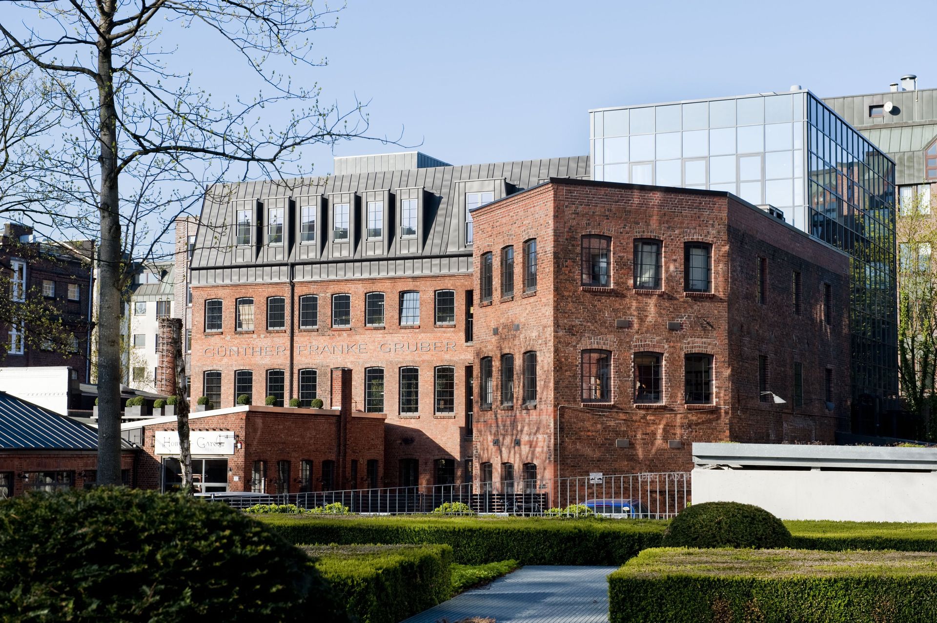 Ein Backsteingebäude mit Bogenfenstern neben einem modernen Glasbüro, eingerahmt von Hecken und kahlen Bäumen