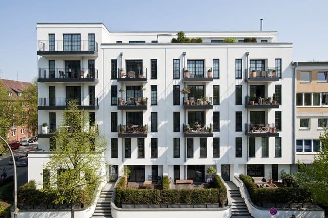 Modernes weißes Apartmentgebäude mit schwarzen Balkonen und grünen Sträuchern davor