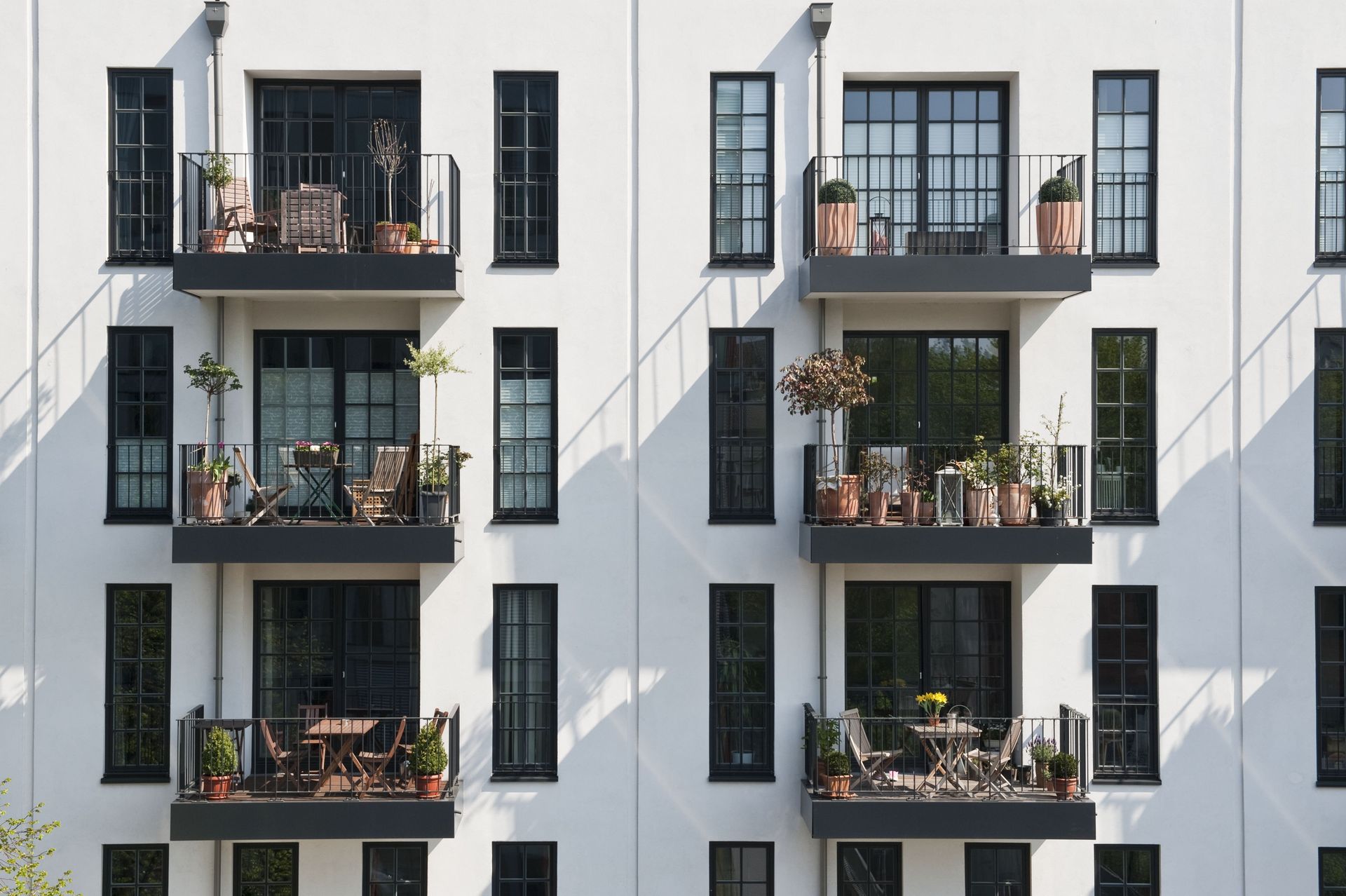 Weißes Wohnhaus mit Balkonen und schwarzen Fensterläden, einige mit Topfpflanzen und Stühlen dekoriert.