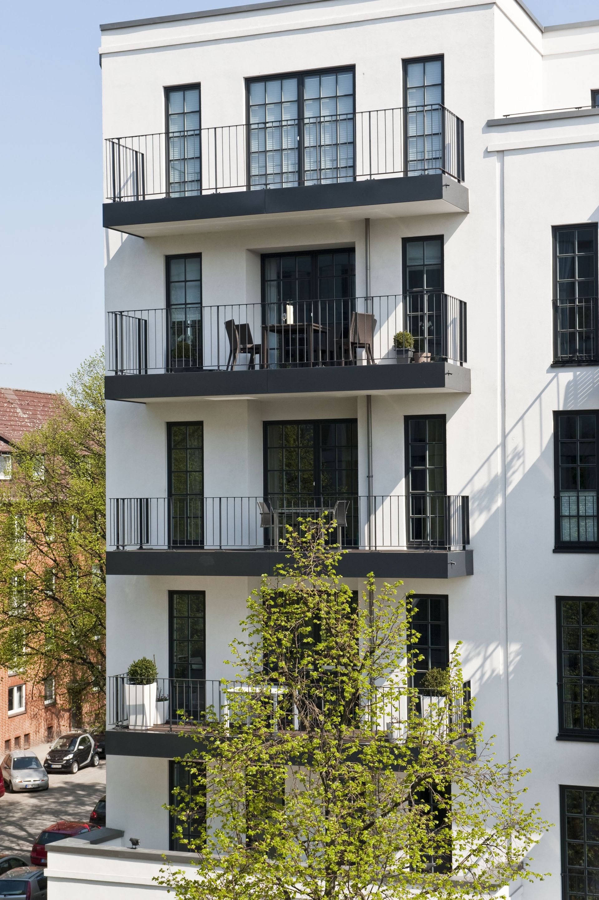 Weißes Wohnhaus mit dunklen Balkonen und großen Fenstern, die teilweise von einem belaubten Baum verdeckt werden.