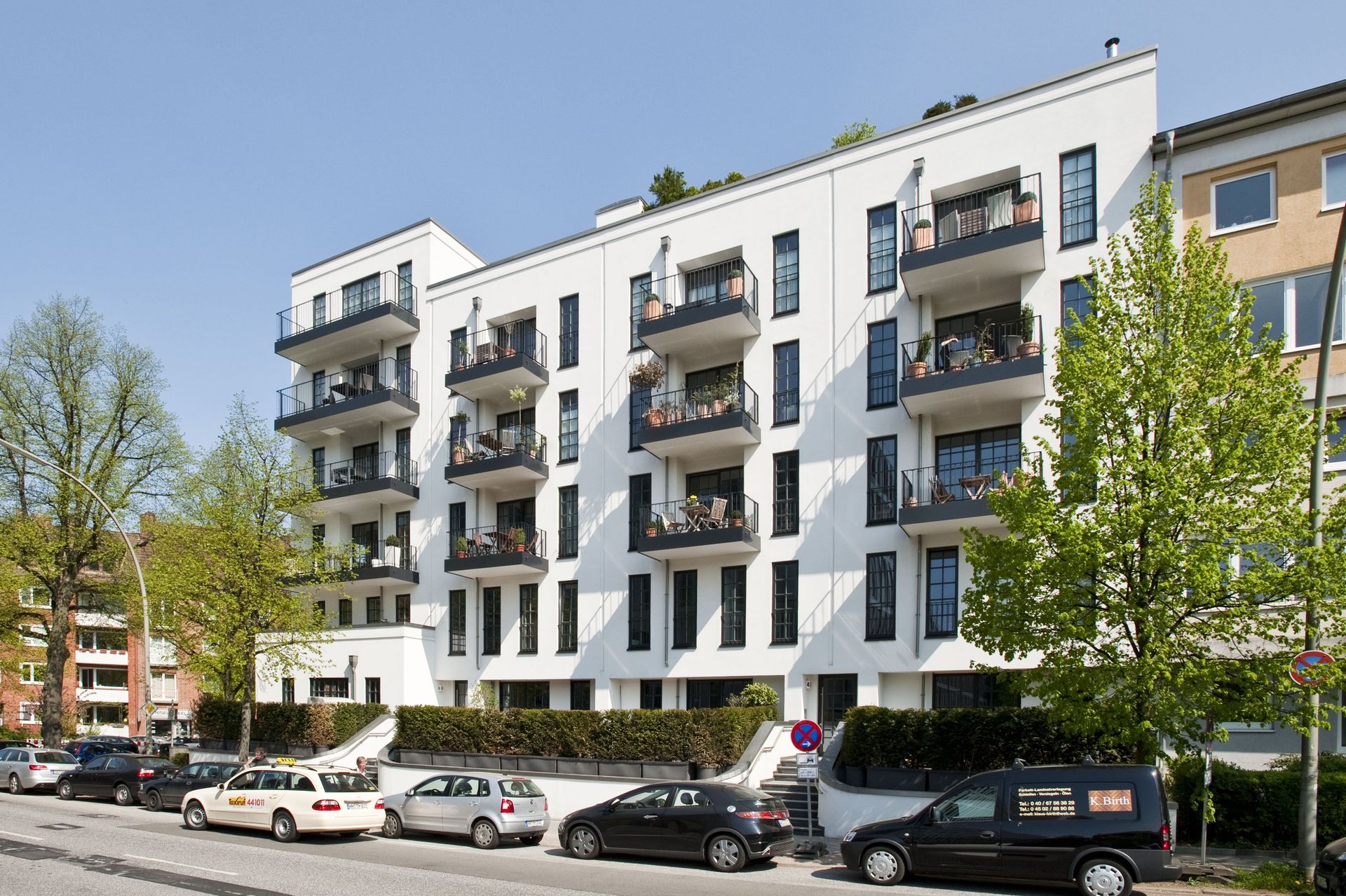 Modernes weißes Wohnhaus mit Balkonen, geparkten Autos und Bäumen entlang einer Stadtstraße