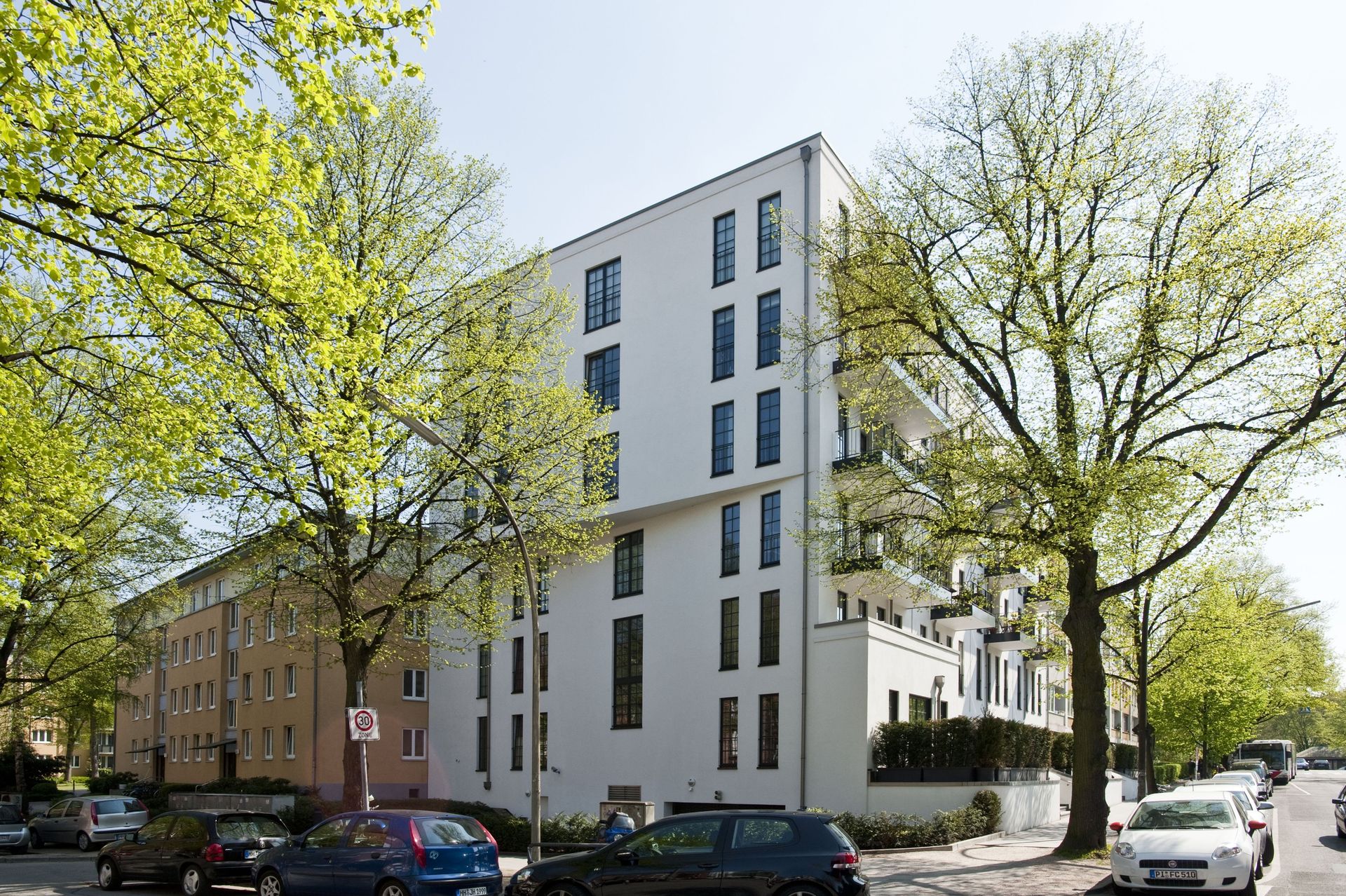Hohes, weißes Wohnhaus an einer von Bäumen gesäumten Straße mit geparkten Autos und sonnigem Himmel