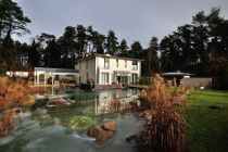 Modernes Haus an einem Teich in der Abenddämmerung, mit Bäumen, Lichtern und einem angelegten Garten.