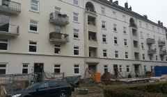 Weißes, mehrstöckiges Wohnhaus mit Balkonen und Fenstern, im Hintergrund einer Baustelle zu sehen.