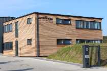 Modernes zweistöckiges Holzhaus mit blau umrandeten Fenstern und einem schrägen, grasbewachsenen Eingang.