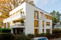 Modernes weißes Mehrfamilienhaus mit Balkonen und Holzfensterrahmen, umgeben von Bäumen.