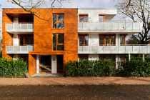 Modernes Wohnhaus mit orangefarbenen und weißen Balkonen, Bäumen davor und einer braunen Fassade im Erdgeschoss.