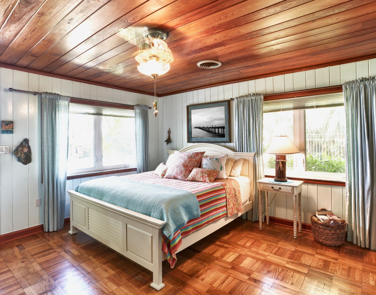 Bedroom with white bed, wood ceiling and floor, light blue curtains, and artwork.
