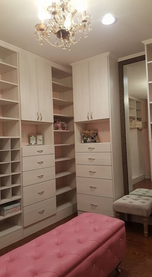 Walk-in closet with cream-colored shelving, drawers, and a pink velvet bench. A chandelier hangs from the ceiling.