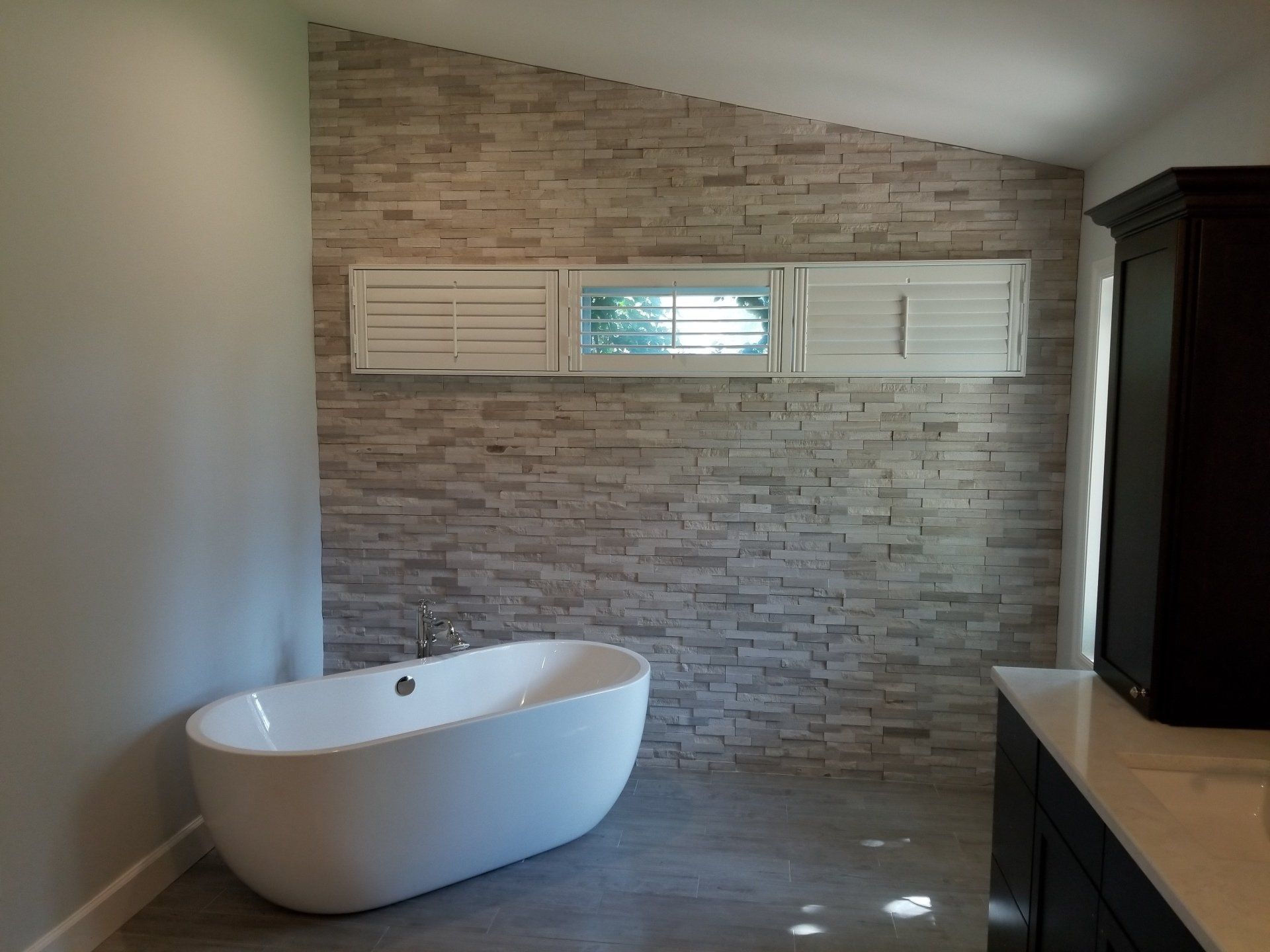 Freestanding white tub in a modern bathroom with stone accent wall, shuttered window, and dark cabinetry.